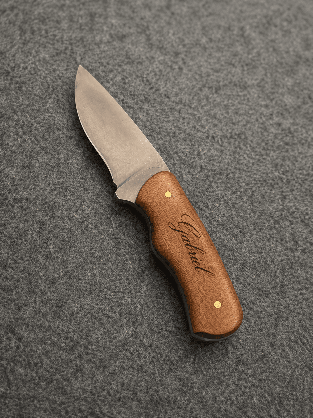 Steel drop-point knife featuring a wooden handle engraved "Gabriel" resting on a textured grey surface.
