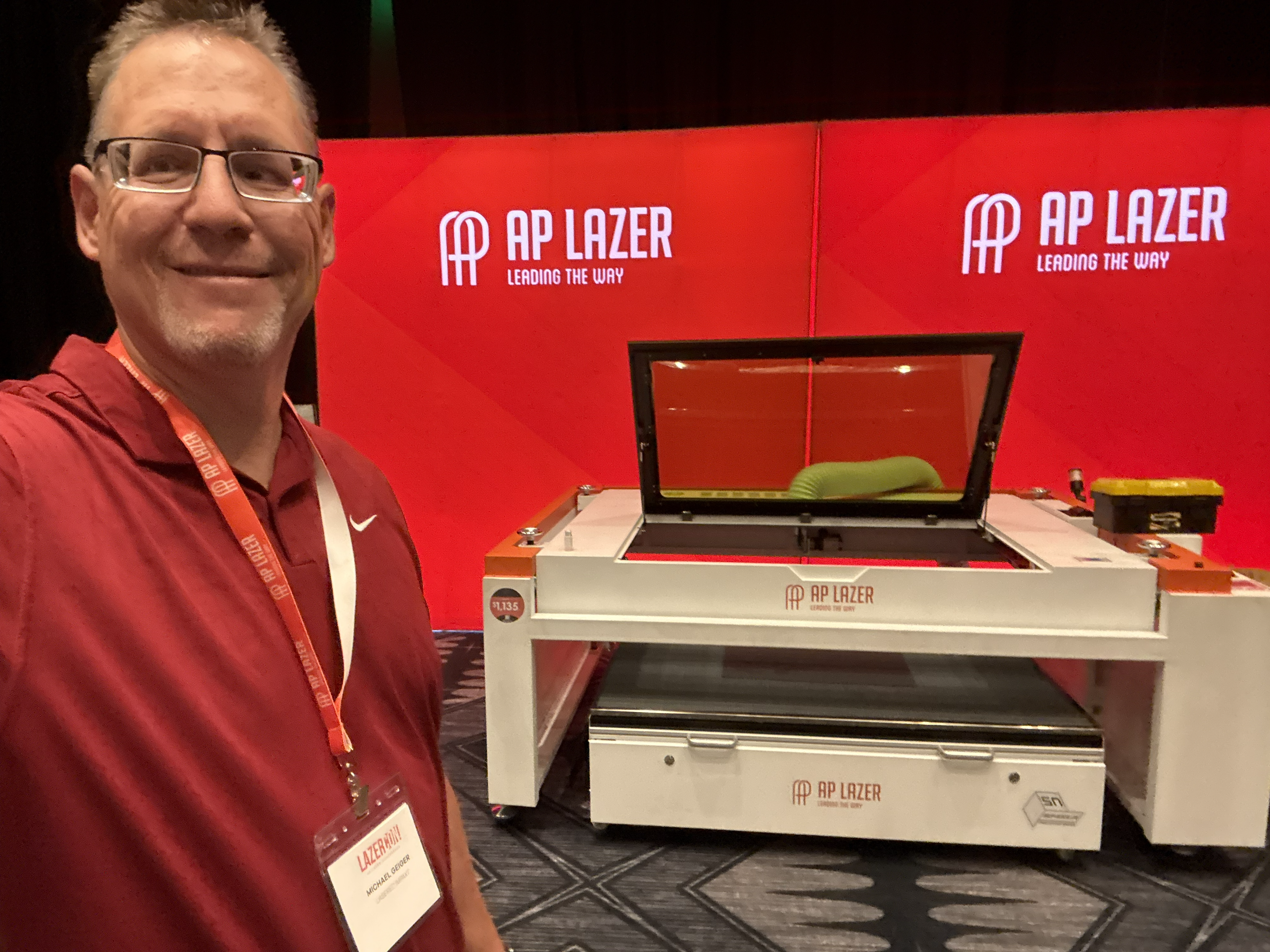 Smiling man takes a selfie with a white AP Lazer machine and red company banners.
